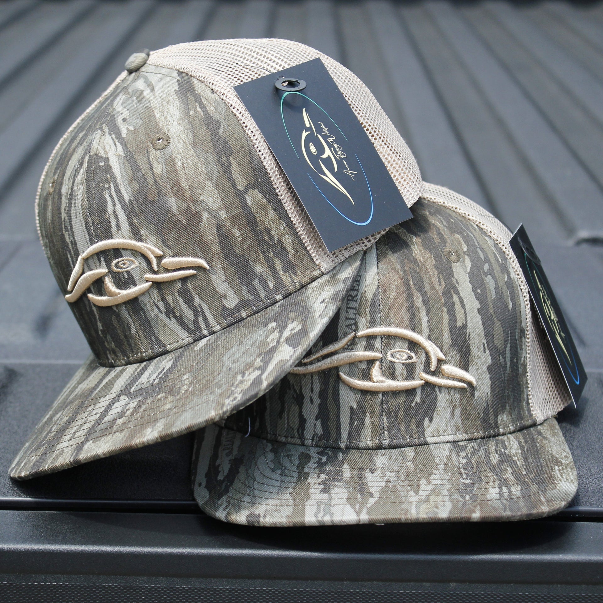 Realtree Legacy AFW Duck Logo Trucker Hat — front view of two camouflage trucker hats with tags on a metallic surface