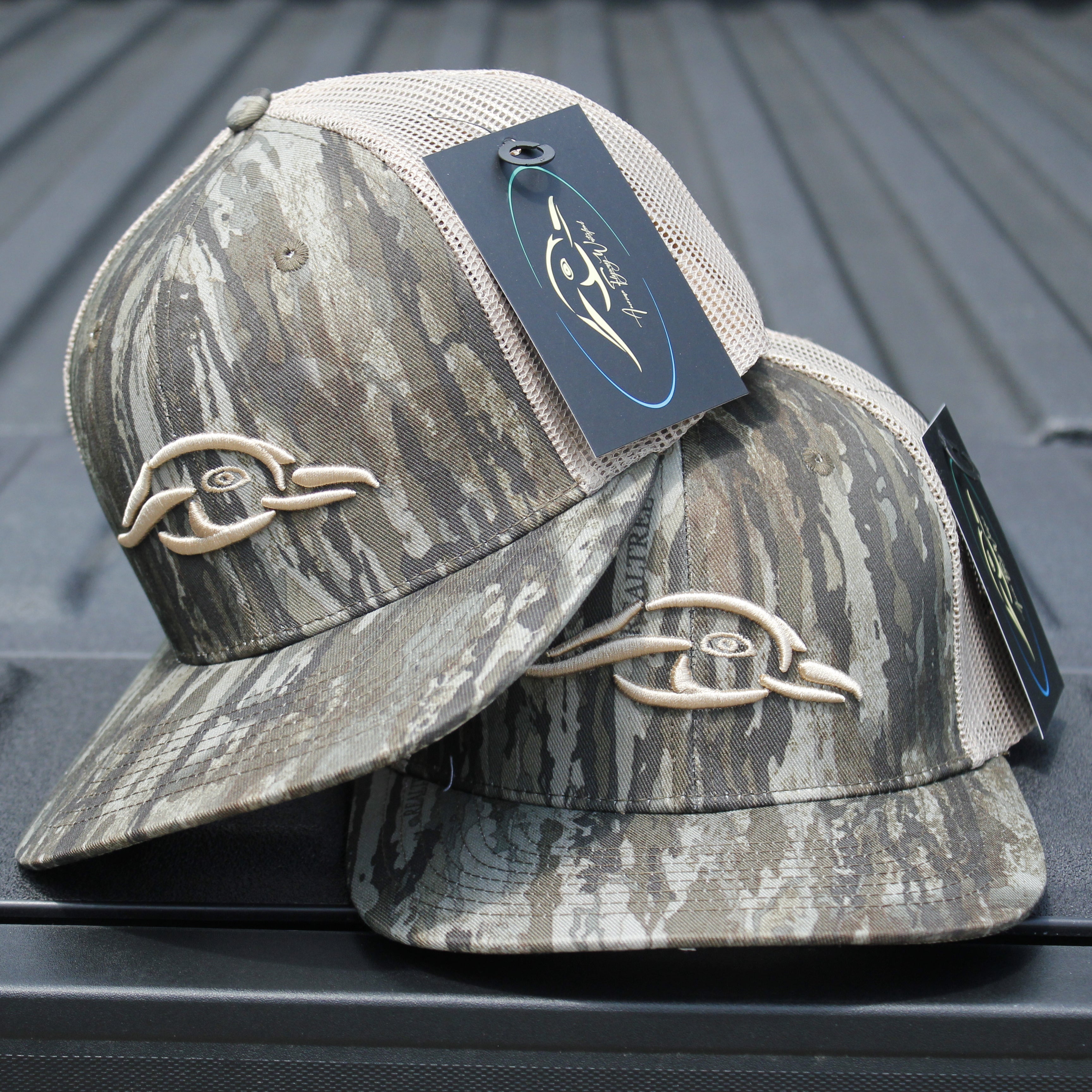 Realtree Legacy AFW Duck Logo Trucker Hat — front view of two camouflage trucker hats with tags on a metallic surface