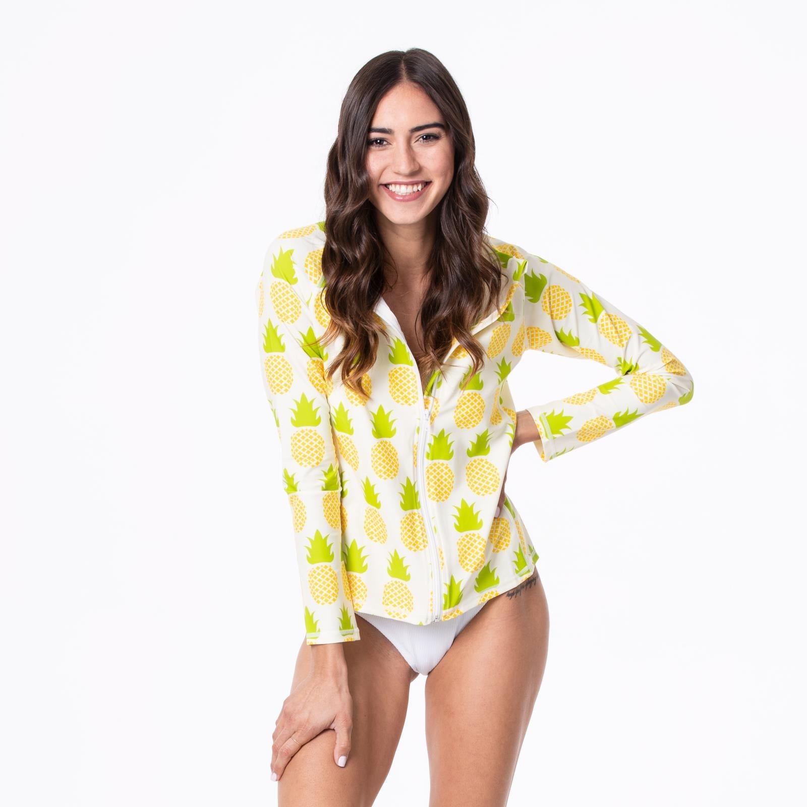 Woman wearing a fitted long sleeve swim cover up in a white and yellow pineapple print with a zip-up collared neckline.