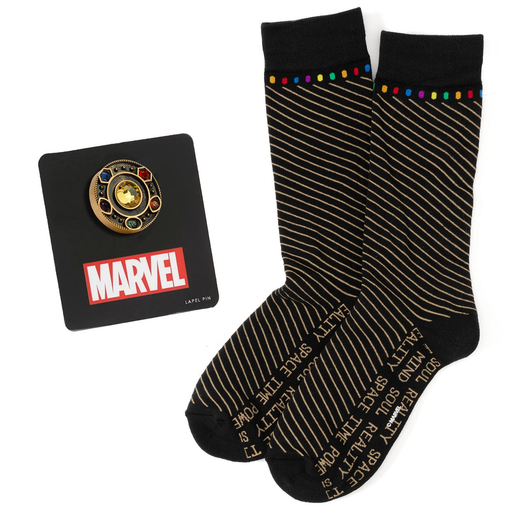 A pair of black crew socks with curved stripes and colorful Infinity Stone accents, displayed alongside an antique gold-tone Avengers lapel pin.