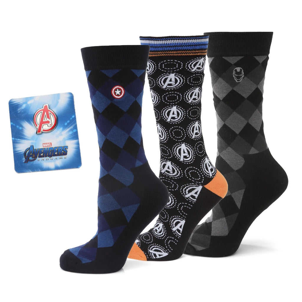 Three pairs of Marvel themed crew socks in blue, black, and grey with argyle and logo patterns next to an Avengers lapel pin in a gift box.