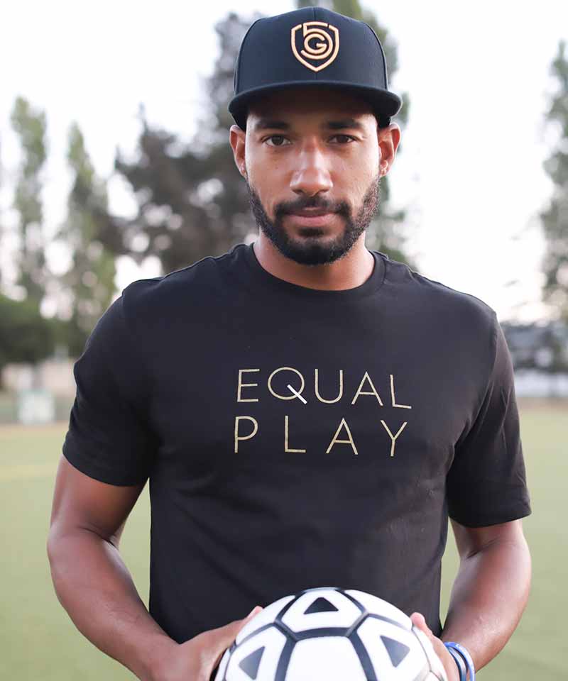 A man wearing a black short-sleeve t-shirt with the words EQUAL PLAY printed on the front, holding a soccer ball.