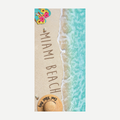 A rectangular beach towel featuring a photographic print of a sandy Miami beach, turquoise water, flip-flops with hibiscus flowers, and a straw hat.