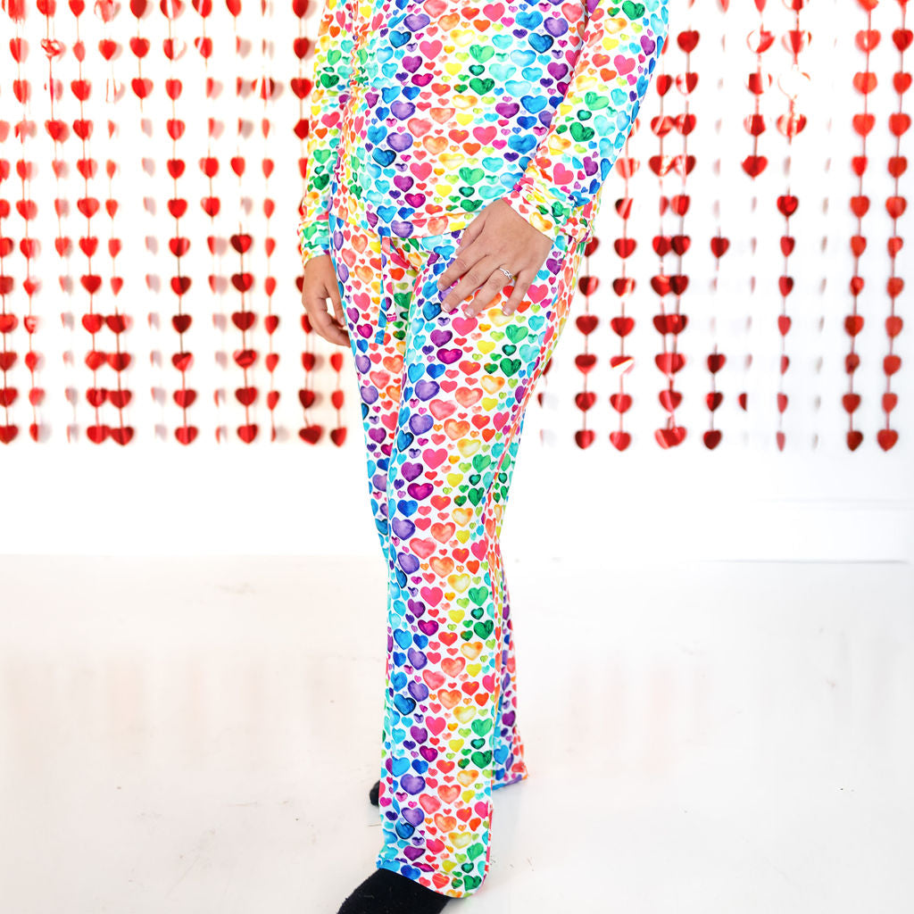 A woman wearing a long-sleeved pajama top and matching pants featuring a colorful rainbow hearts print against a heart-themed backdrop.