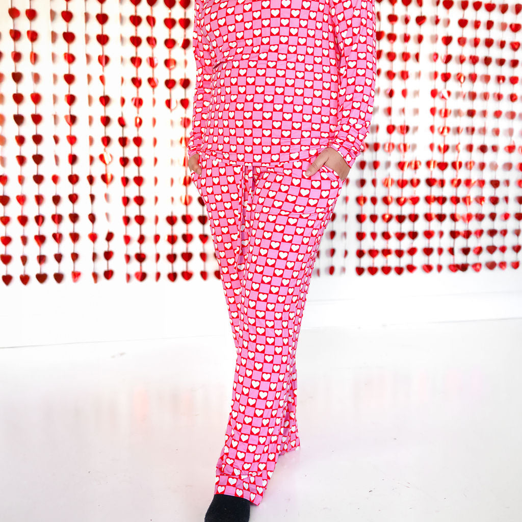 A woman wearing a matching two-piece pajama set with a pink and red checkerboard heart pattern, featuring a button-front top and pants with pockets.
