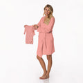 A woman modeling a blush colored maternity lounge robe while holding a matching patterned baby gown and hat in Blush Stork print