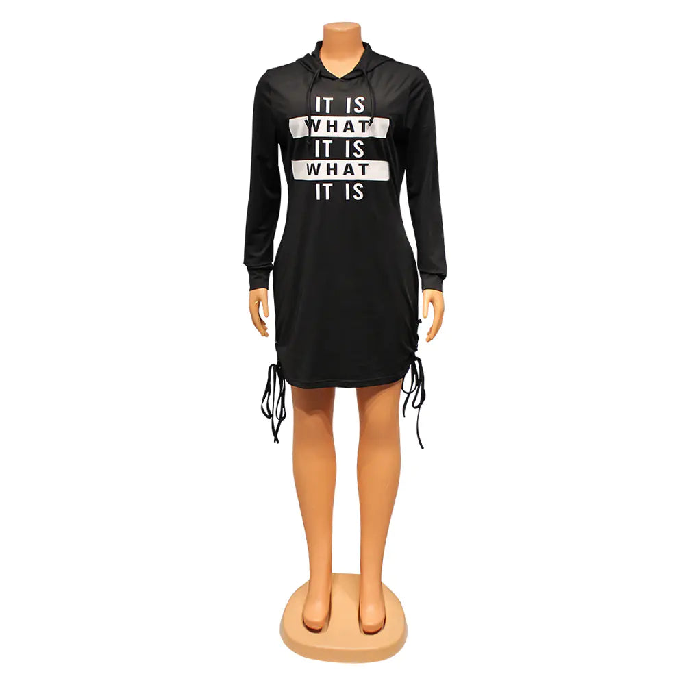A woman wearing a black long-sleeved mini hooded t-shirt dress with white 'It Is What It Is' graphic text and side drawstring lace-up details.