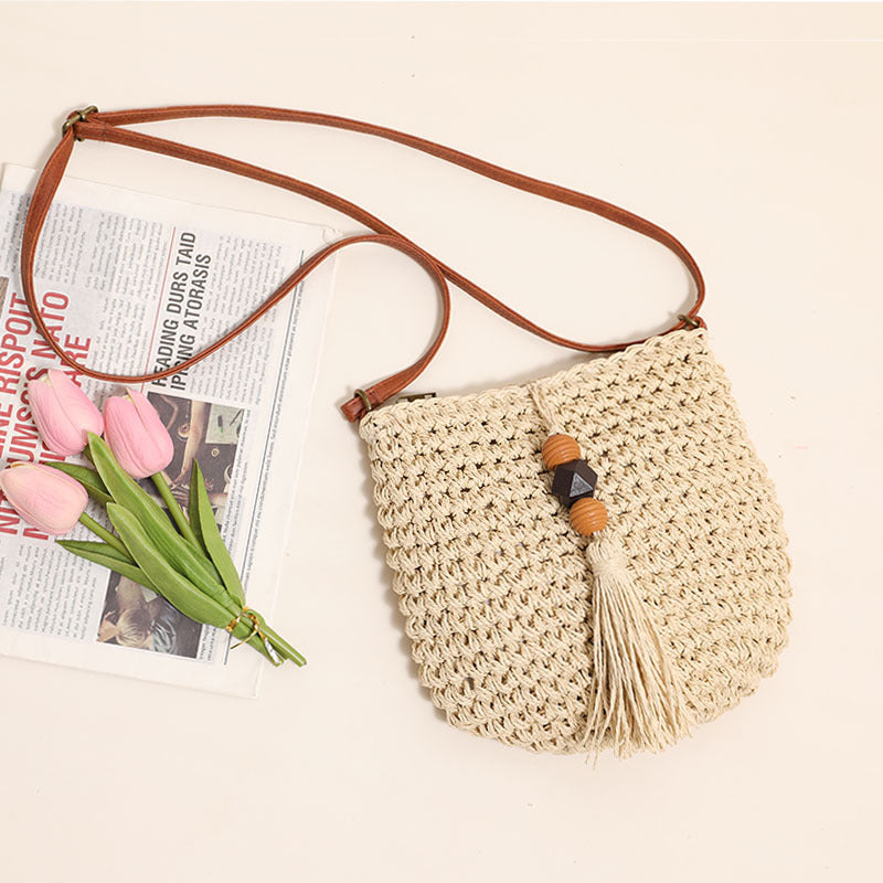 Woven straw crossbody bag featuring wooden beads and a decorative tassel with an adjustable brown strap on a light background