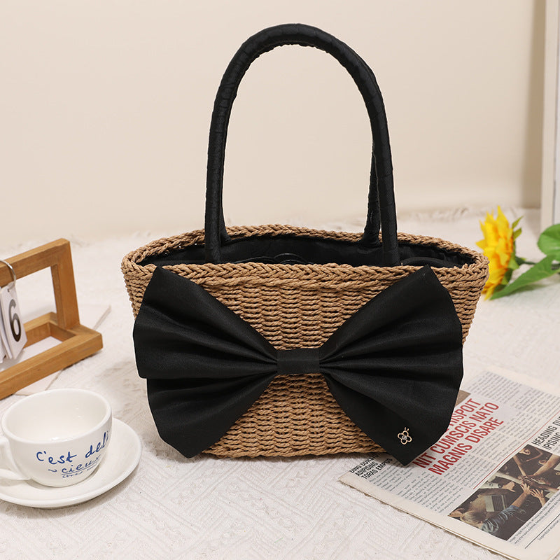 A tan woven straw tote bag featuring a large black fabric bow on the front, displayed on a neutral surface.