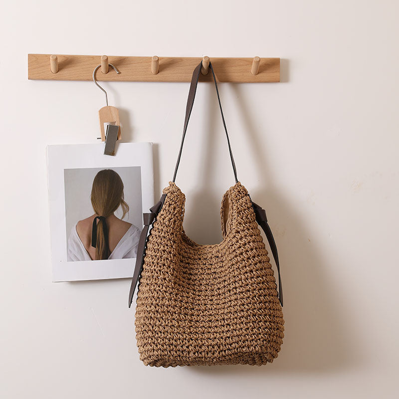 A beige woven straw shoulder bag with dark brown straps and side tassel detail, displayed against a neutral background.