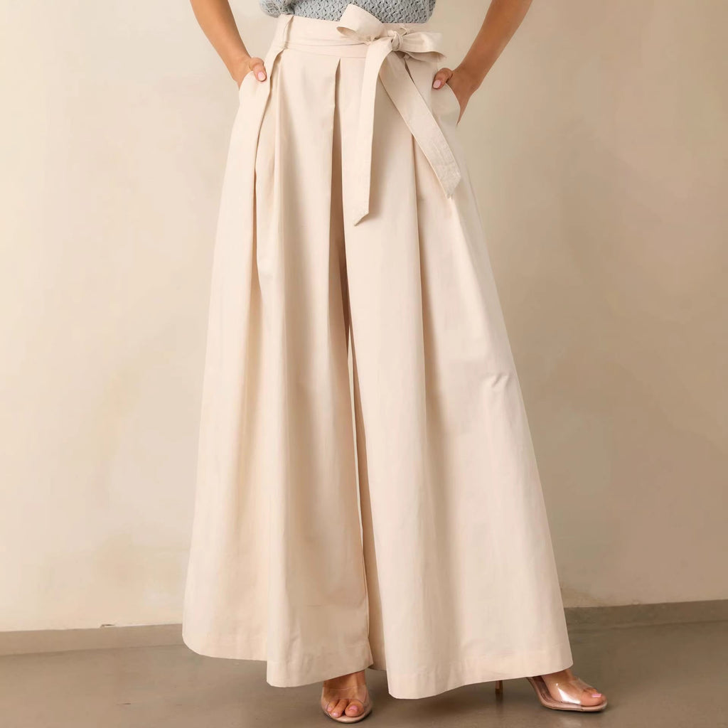 A model wearing Bella Mia women's apricot high-rise belted pleated wide-leg pants with a light blue top and clear heels, holding a matching handbag.