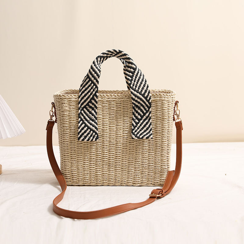 Square woven straw crossbody bag with brown faux-leather strap and a monochrome geometric pattern scarf wrapped around the top handles.
