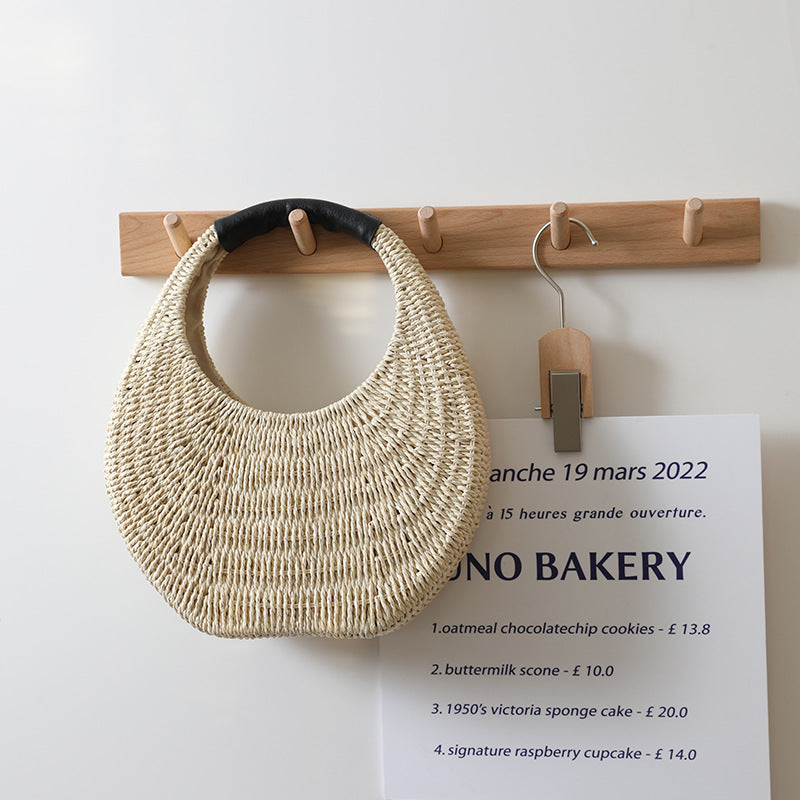 Crescent-shaped natural woven straw handbag with a black leather handle resting on a white surface.