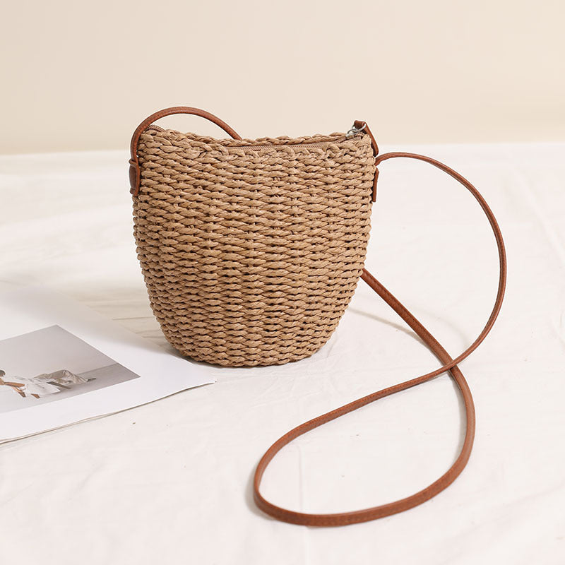 Simple woven straw bucket bag with a thin brown shoulder strap on a white background