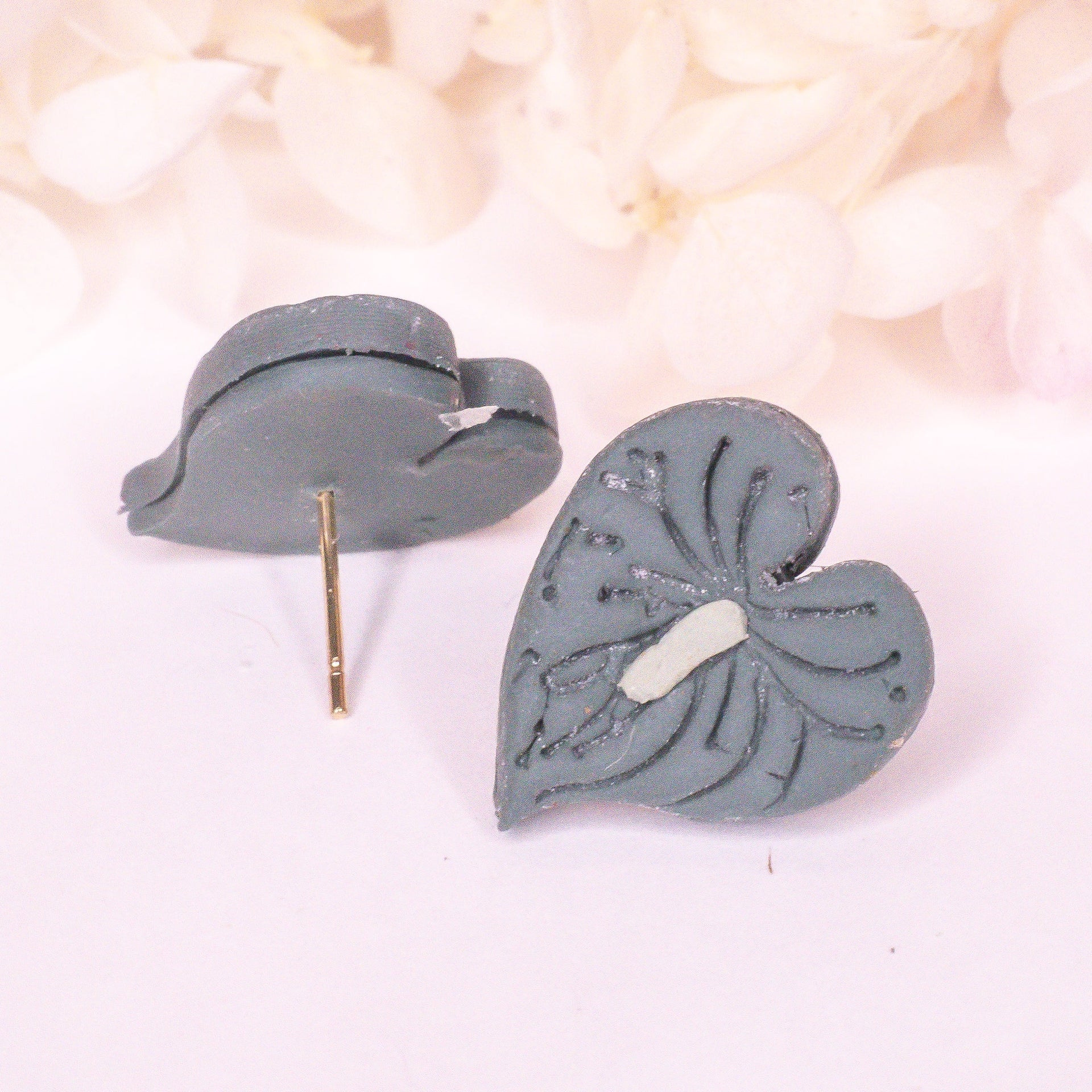 Close up of mini green heart-shaped anthurium stud earrings with engraved texture on a neutral background.