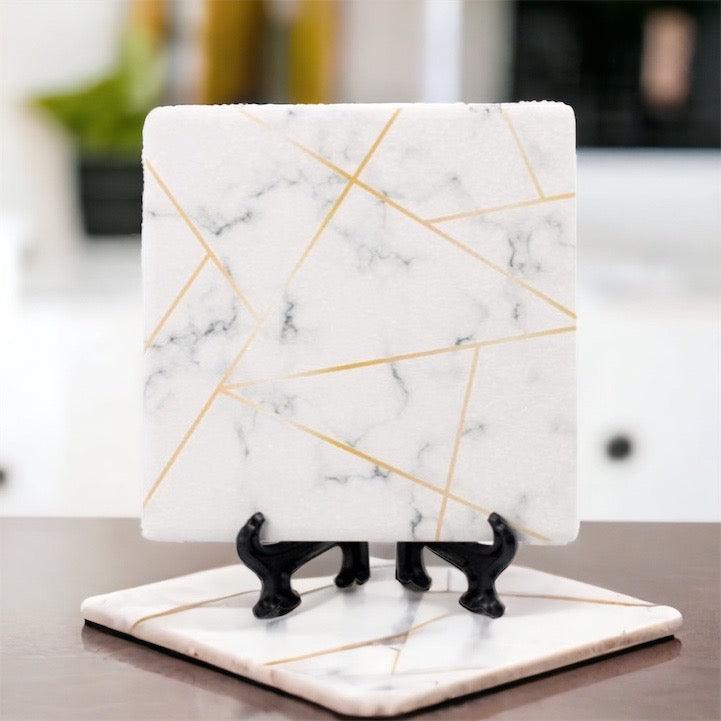 Set of square ceramic coasters with white marble pattern and gold geometric lines on a stand