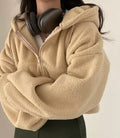 A woman wearing a cozy, fuzzy beige sherpa zip-up hoodie with a hood up, styled for a casual look.