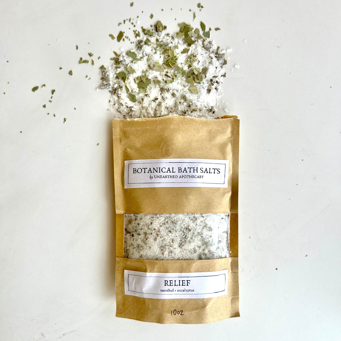 A 10oz pouch of Relief botanical bath salts with menthol and eucalyptus, shown next to a sprig of fresh eucalyptus.