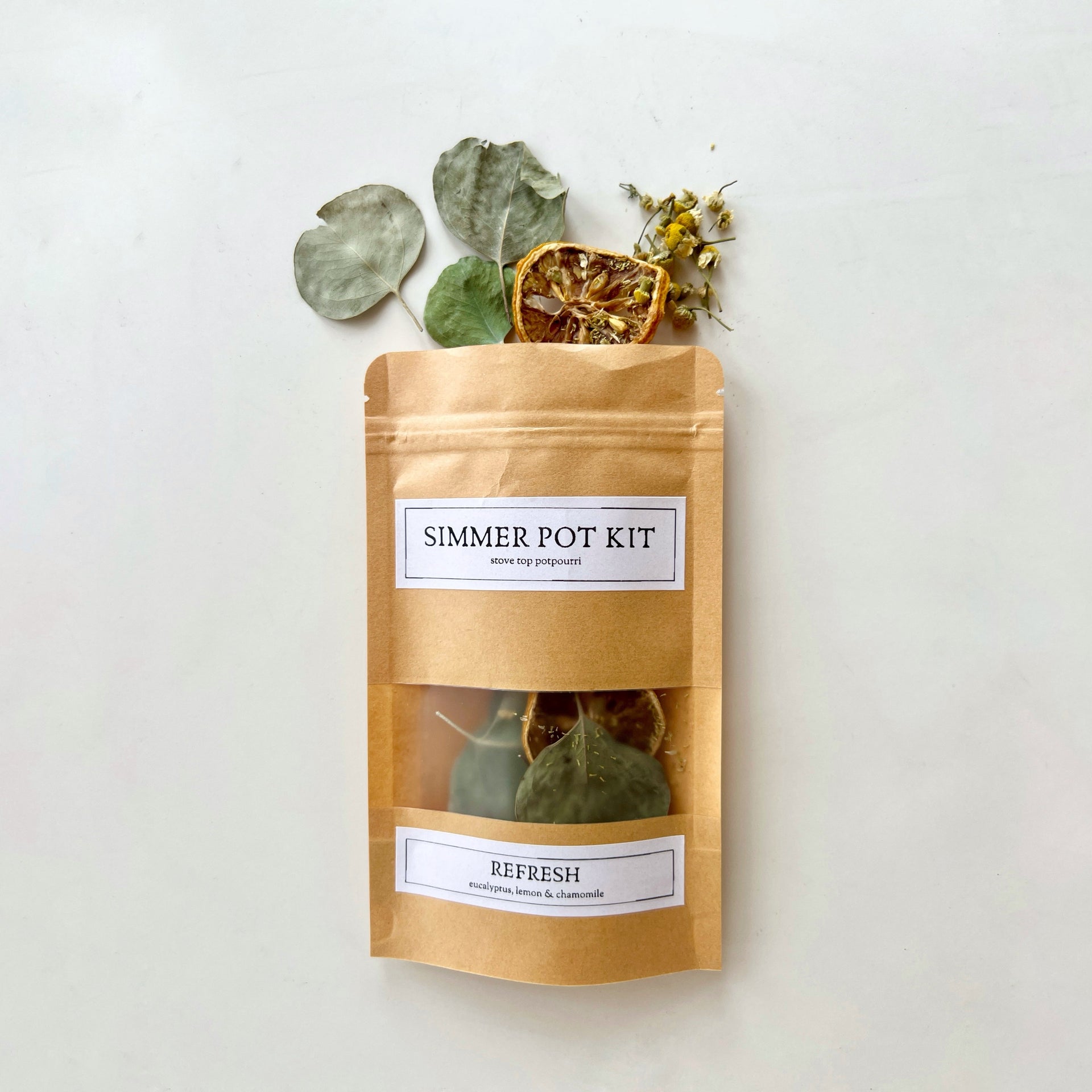 A paper pouch of Refresh Simmer Pot Kit containing dried eucalyptus, lemon slices, and chamomile flowers, with loose ingredients scattered next to it.