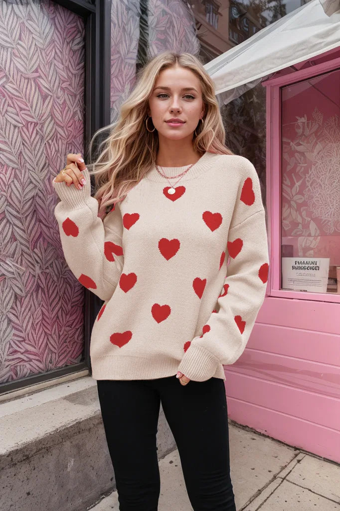 A woman wearing a beige knit sweater covered in a red heart pattern with a crew neckline and long sleeves.