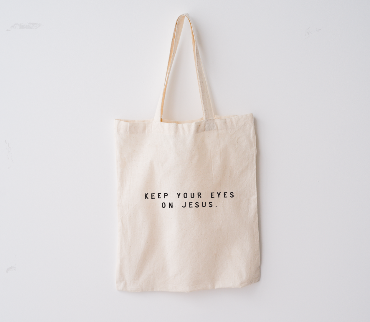 Keep Your Eyes On Jesus canvas market tote in beige with black text, shown on a neutral background.