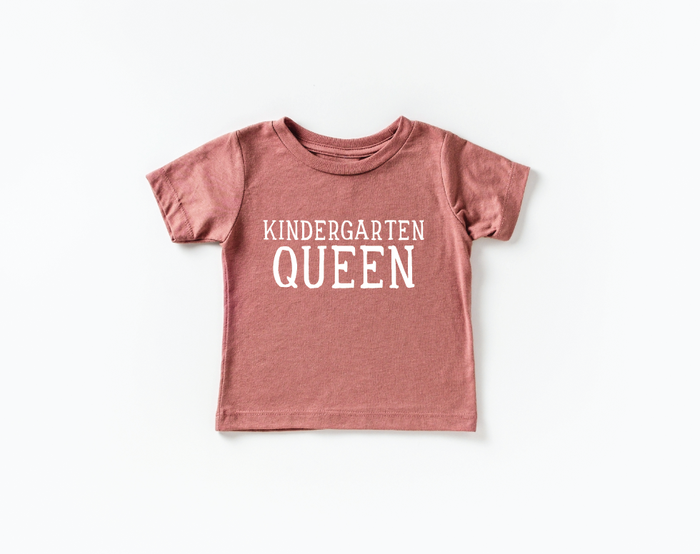 Little girl wearing a Kindergarten Queen graphic tee in dusty-rose