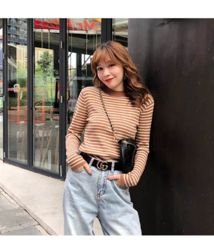 A woman wearing a tan and white striped mock neck long sleeve ribbed sweater with a belt and blue jeans.
