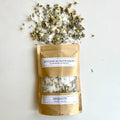 A kraft paper pouch of Serenity botanical bath salts spilled out showing white salt crystals, dried lavender buds, and whole chamomile flowers.