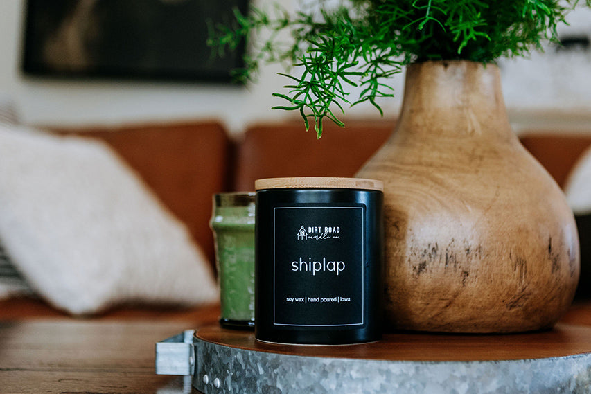 A black tin candle labeled Shiplap with a wood lid sitting on a rustic galvanized tray.