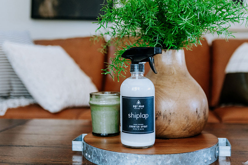 A clear glass spray bottle with a black label labeled Shiplap Counter Spray, sitting on a wooden and metal tray.