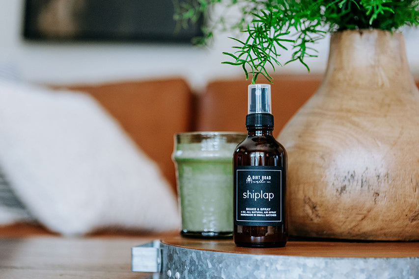 Amber glass bottle of Shiplap air and room spray sitting on a wooden surface next to a plant.