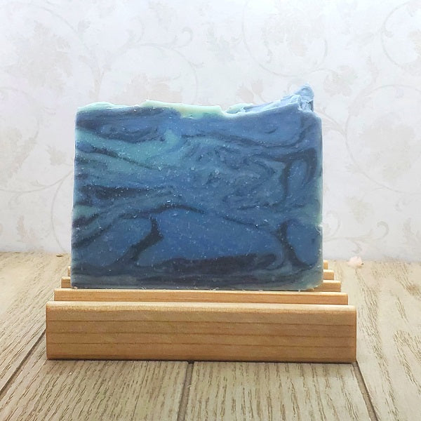 A rectangular bar of blue and navy swirled soap resting on a light wooden soap dish against a floral wallpaper background.