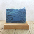 A rectangular bar of blue and navy swirled soap resting on a light wooden soap dish against a floral wallpaper background.