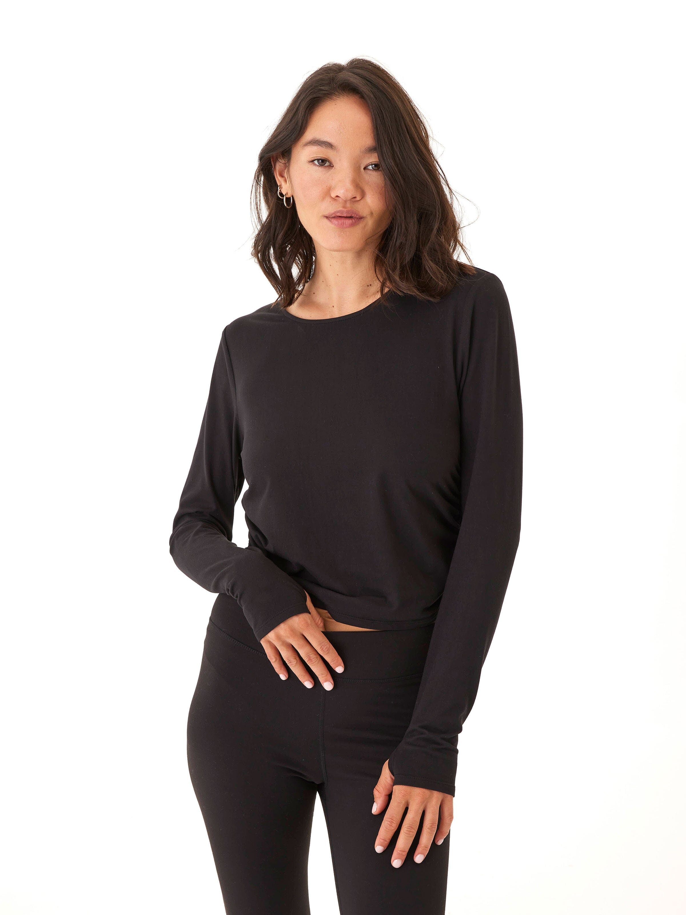 A woman wearing a fitted black cropped long sleeve top with ruched sides and thumbholes, styled for an athletic aesthetic.