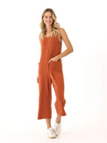 A woman wearing the sleeveless Rainey Patch Pocket Jumpsuit in terracotta with a wide-leg ankle fit and patch pockets.