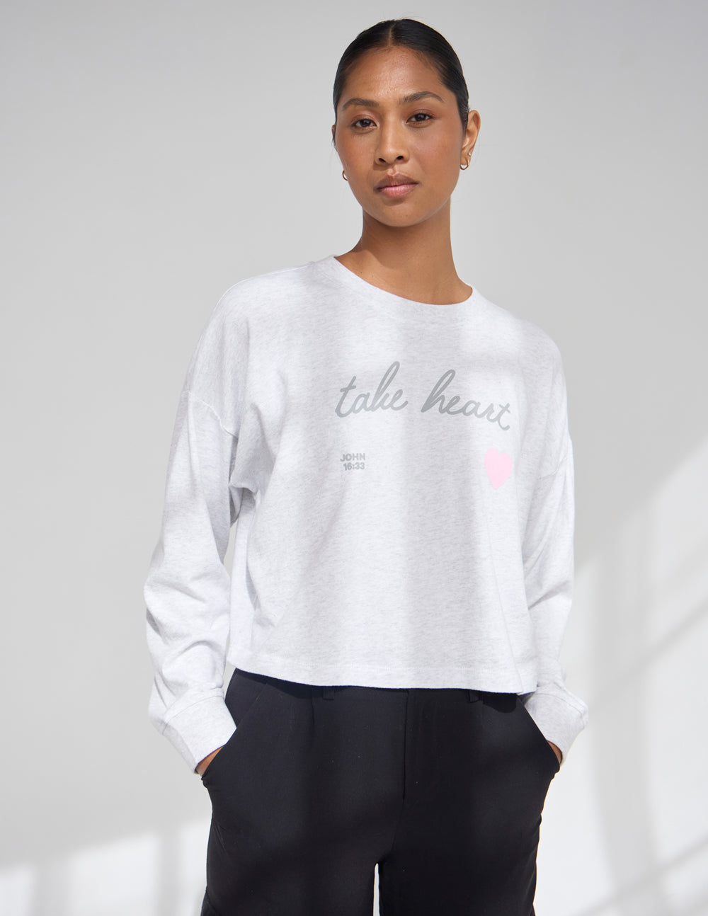 Woman wearing a light grey heather cropped long sleeve tee with soft pink Take Heart text, featuring a relaxed fit and crew neckline.