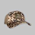 Tan woven duck camo trucker hat featuring a detailed patch and mesh back paneling.