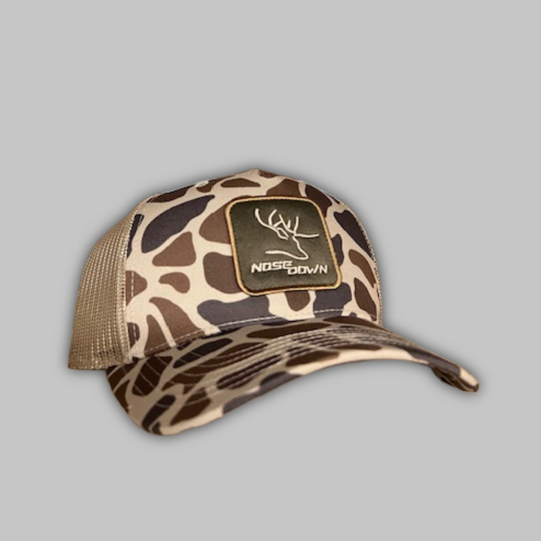 Tan woven duck camo trucker hat featuring a detailed patch and mesh back paneling.