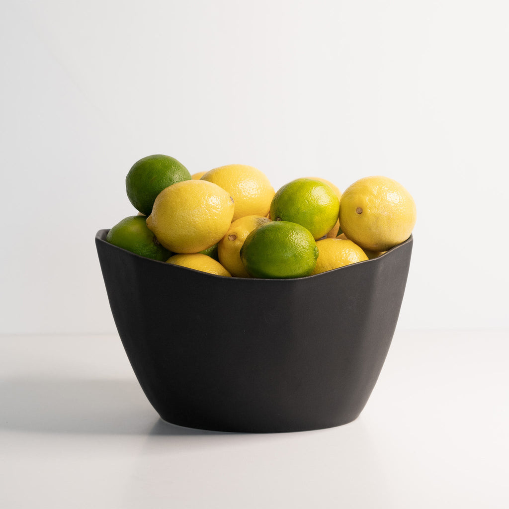 A medium-sized matte black porcelain bowl filled with fresh lemons and limes, against a plain white background.