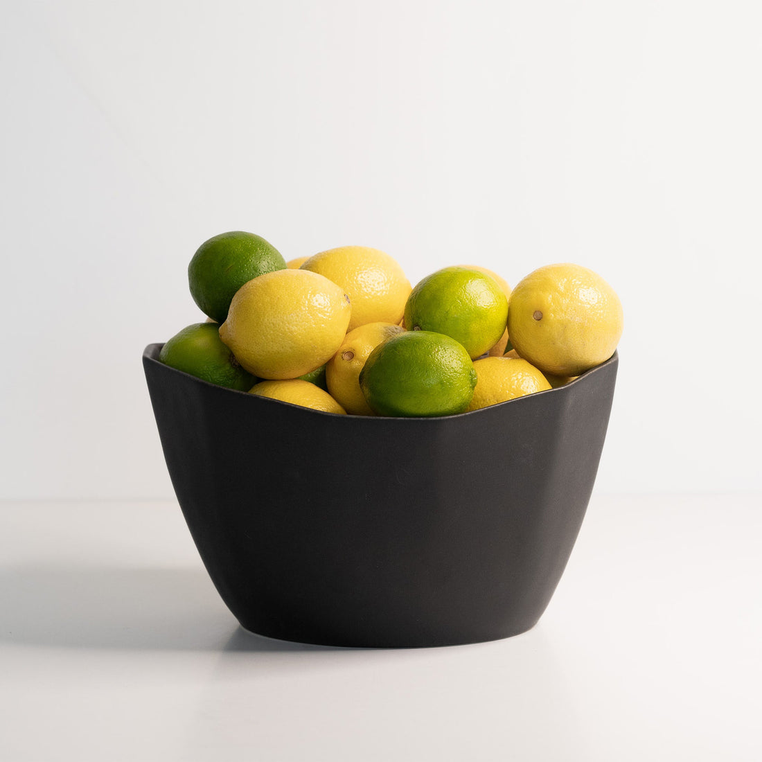 A medium-sized matte black porcelain bowl filled with fresh lemons and limes, against a plain white background.