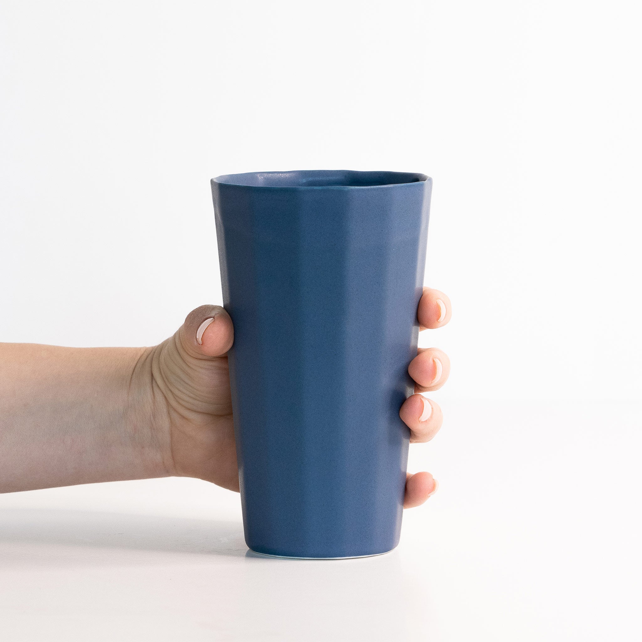 A hand holding a minimalist blue matte porcelain pint cup against a white background