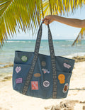 Large dusty blue denim tote bag with grey straps and assorted iron-on patches displayed on a flat surface.