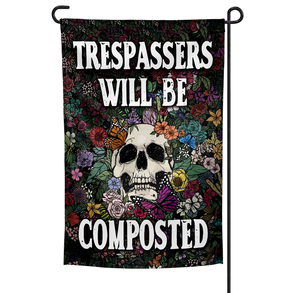 Trespassers Will Be Composted garden flag in black and multi-color displayed on a metal stake in a garden setting.