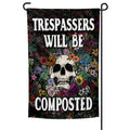 Trespassers Will Be Composted garden flag in black and multi-color displayed on a metal stake in a garden setting.