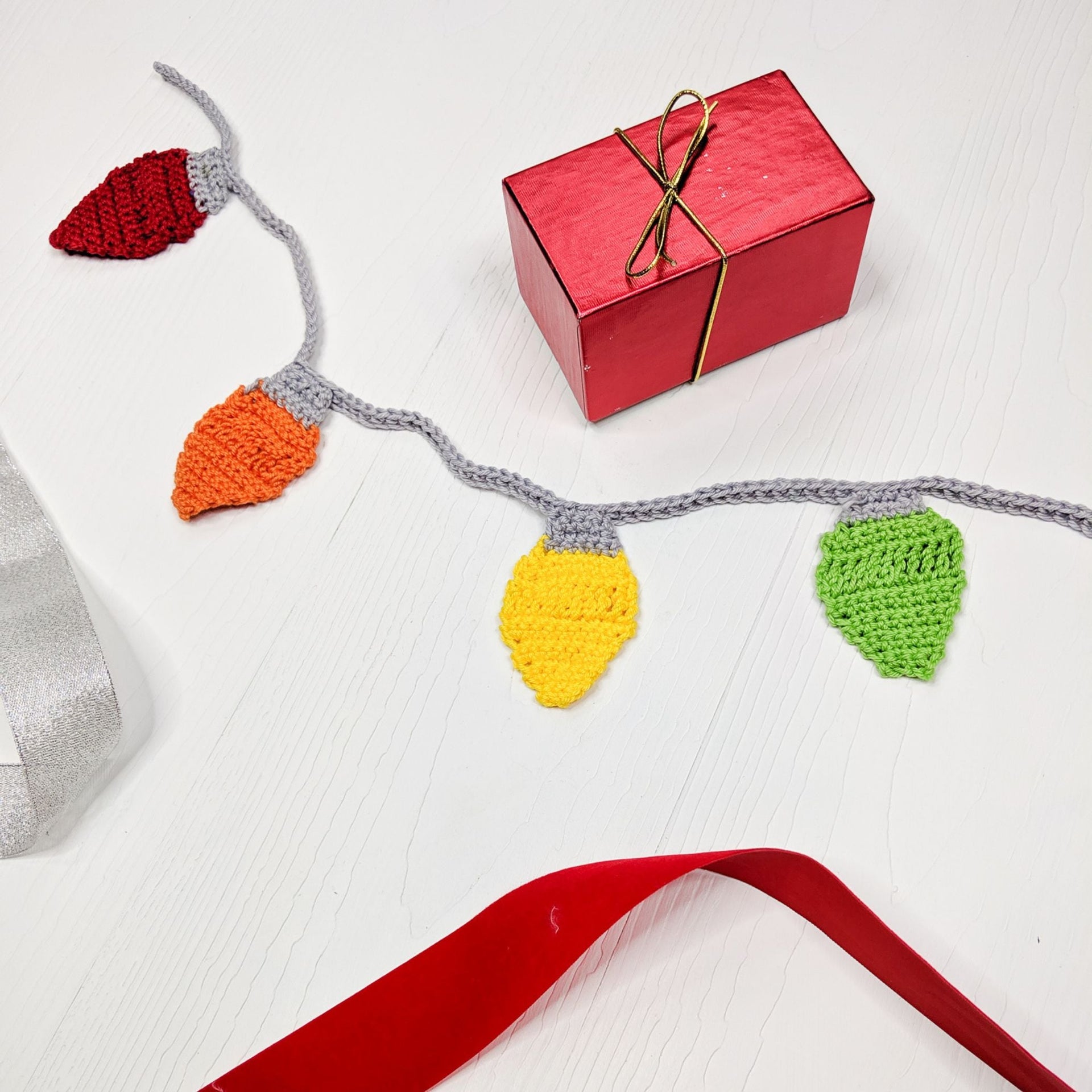 Colorful crochet Christmas light bulb garland made with red, orange, yellow, green, and grey knit yarn draped on a wooden surface.