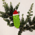 A handmade green, red, and white crocheted Christmas pickle ornament hanging on a festive tree branch.