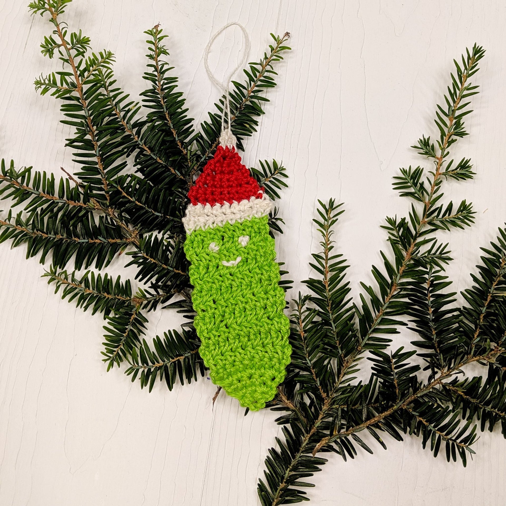 A handmade green, red, and white crocheted Christmas pickle ornament hanging on a festive tree branch.