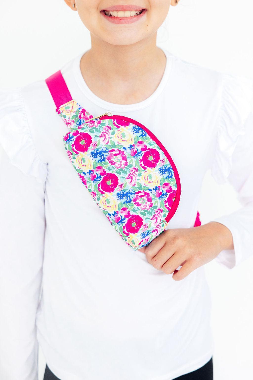 A young girl wearing a bright floral patterned belt bag across her chest, featuring pink, yellow, and blue flowers.