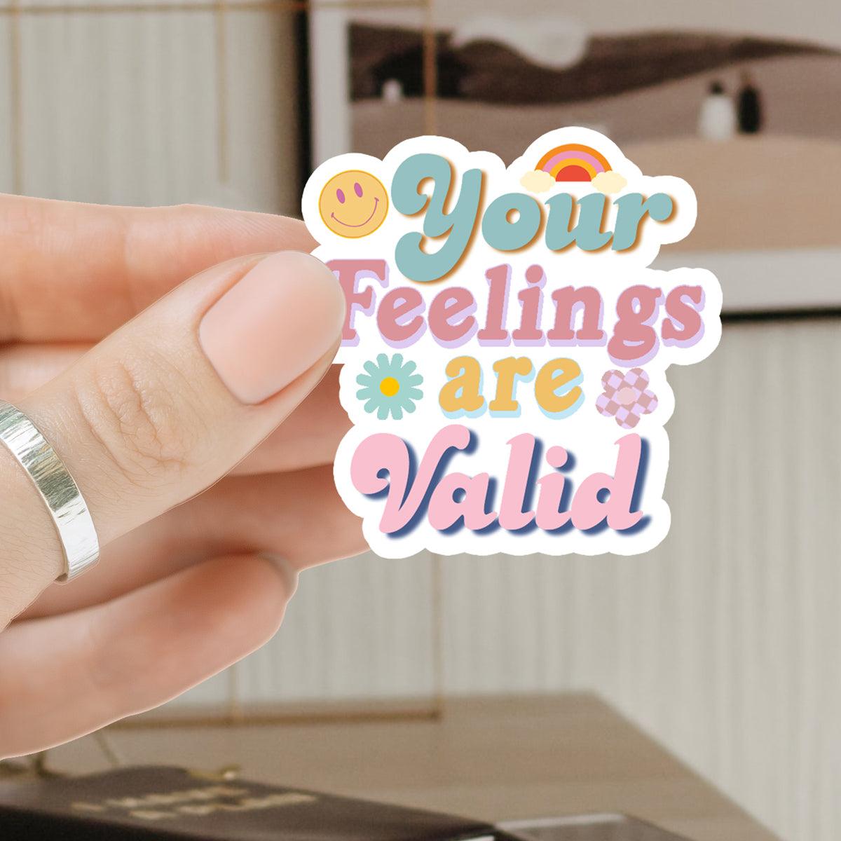 A vinyl sticker with the text Your Feelings Are Valid in retro typography, surrounded by a smiley face, rainbow, and small flowers, held against a black journal.