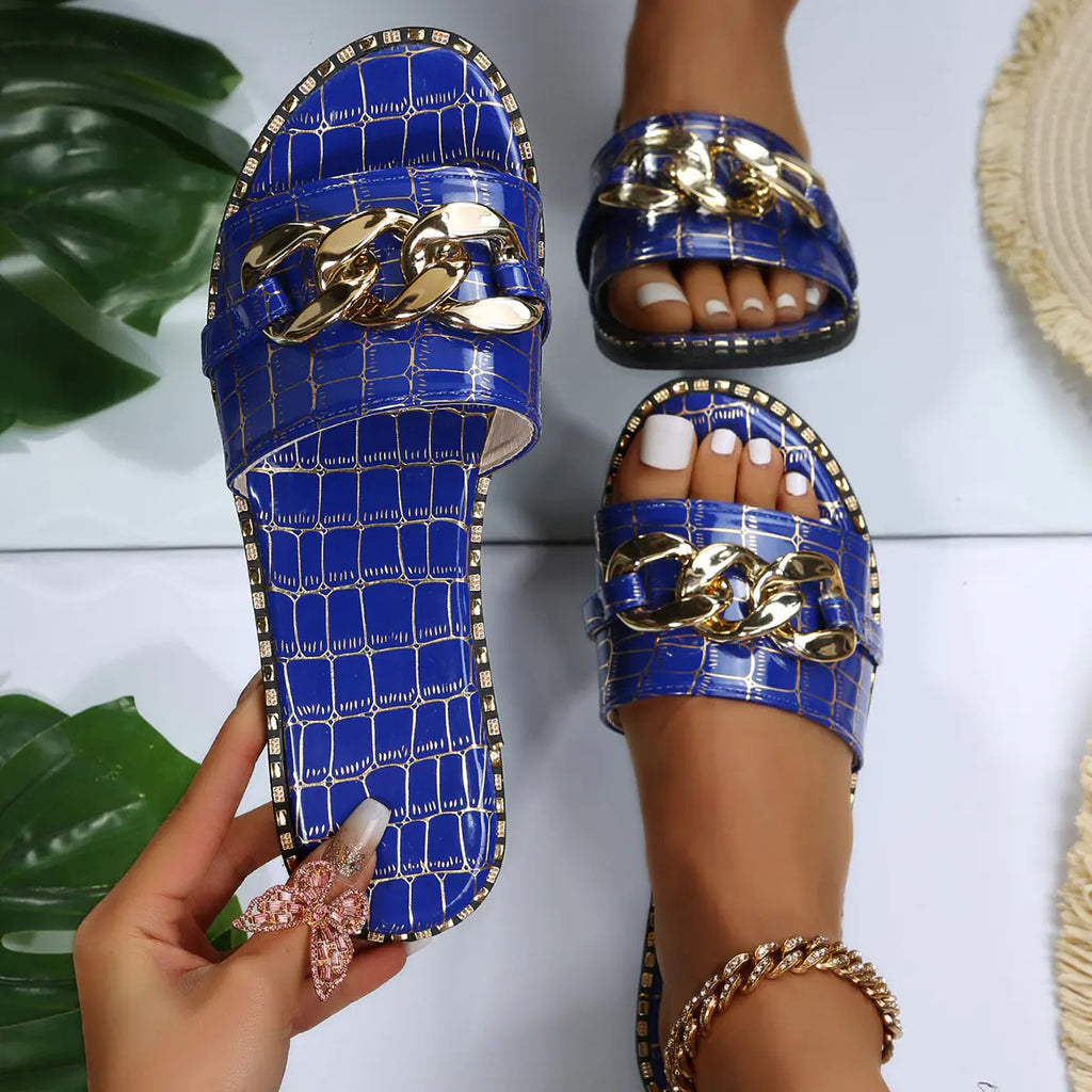 A pair of bright blue croc-embossed flat slide sandals featuring a decorative chunky gold chain accent on the strap.